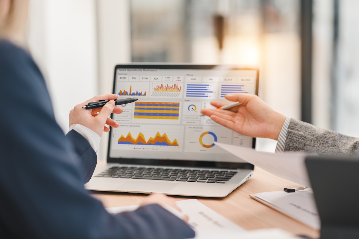 Two people review charts and graphs on a laptop screen, pointing to performance data during a discussion.