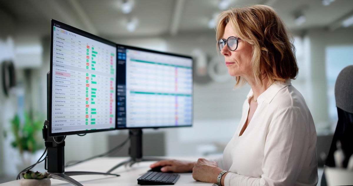 A professional works at a desk in a modern office, viewing spreadsheets on dual monitors.
