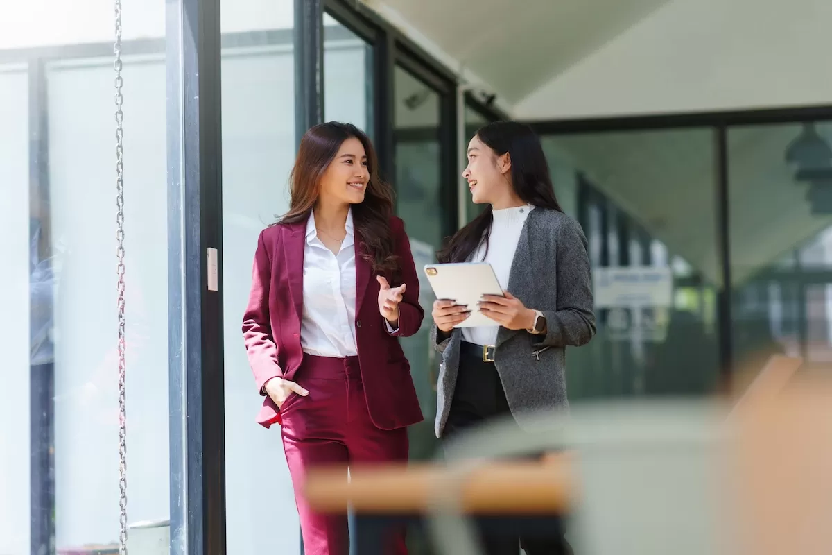 Two professionals walking and talking in a modern office hallway, one holding a tablet while they discuss work
